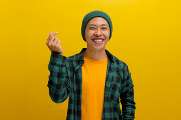 Attractive young Asian man, dressed in beanie hat and casual shirt, exudes happiness as he creates a romantic mini heart shaped gesture with his finger, expressing tender feelings, isolated on yellow