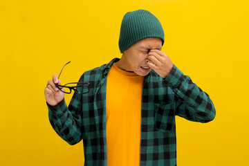 Asian man in a beanie and plaid shirt rubs his irritated eyes, pinching his nose in discomfort. He...
