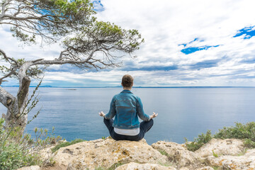 Junge Frau meditiert an einem Felsvorsprung am Meer mit Baum
