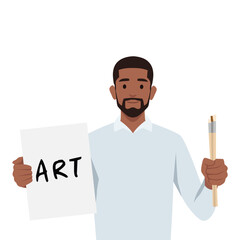 Young man holding paper with art written on it and holding paint brushes ready to paint.