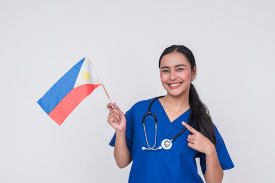 A young Filipino female nurse wearing blue scrubs, pointing to a Philippine flag, her home country.