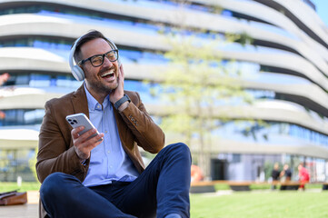 Ecstatic handsome entrepreneur with cellphone laughing and enjoying music over headphones in city