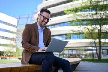 Low angle view of happy young man working online over laptop computer while sitting on bench in city