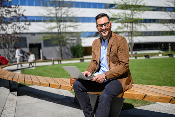 Happy young manager working online over computer while sitting on bench against modern building