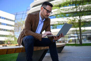Male professional reading e-mails with focus over computer while sitting on bench in modern city