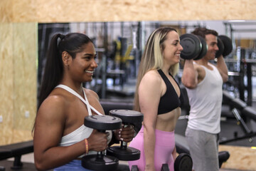 Group of multiracial smiling people exercising their arms with dumbbells in a gym, sport dress, side view. Fitness and healthy lifestyle concept