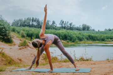 Woman performing a Triangle Pose (Trikonasana) on a yoga mat by a river, surrounded by natural scenery