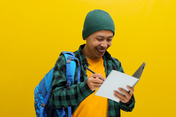 Excited Asian student, dressed in a beanie hat and casual clothes with a backpack, is diligently working on straightforward homework, writing something in his notebook, isolated on yellow background