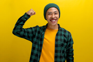 Excited Asian man, dressed in a beanie hat and casual shirt, is making a strong gesture by lifting his arm to show muscles, expressing pride in his achievements, standing against a yellow background