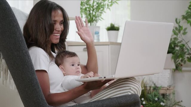 Smiling mixed race woman video conference with friends or relatives holding baby in her arms and waving affably at laptop camera