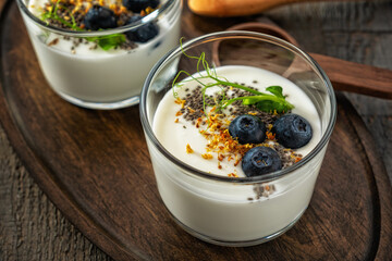 Cups of homemade yogurt on a wooden tray on a textured gray wooden background, decorated with blueberries, chia seeds, dried osmanthus flowers and pea sprouts. Side and top view.