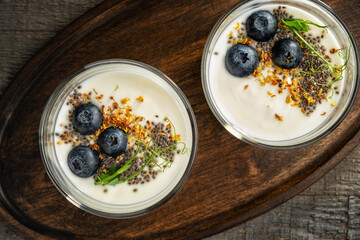 Homemade yogurt in two glass cups on a dark wooden tray garnished with blueberries, chia seeds, dried osmanthus flowers and pea sprouts. View from above. Close-up.