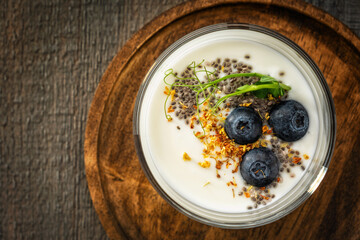 Close-up of homemade yogurt in a glass cup decorated with blueberries, chia seeds, osmanthus flowers and pea sprouts on a round textured wooden plate on a gray wooden background. View from above.