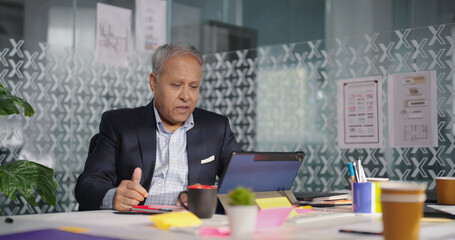 Indian aged older formal dress man expert staff sitting use tablet touch pad talk online video call share ideas plan indoor office old elder busy boss ceo male agent look screen job centre work place