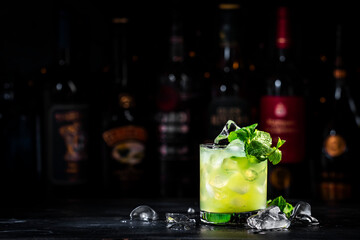 Green Negroni cocktail drink with gin, aperitif, liqueur, mint and ice. Black bar counter background with bottles