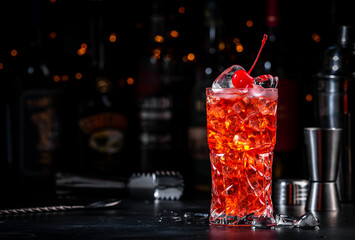 Red cocktail drink with pink vermouth, liqueur, cherry, juice and ice, black bar counter background, steel tools and bottles