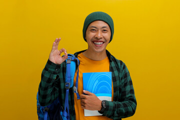 Friendly young Asian student, dressed in a beanie hat and casual shirt and carrying backpack, is smiling at the camera while making the OK sign gesture and holding books, isolated on yellow background