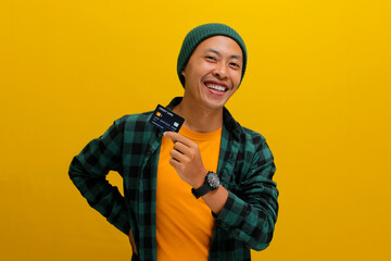 A happy young Asian man, dressed in a beanie hat and casual shirt, enthusiastically shows a credit card in his hand, feeling excited while demonstrating a solution for online shopping payment