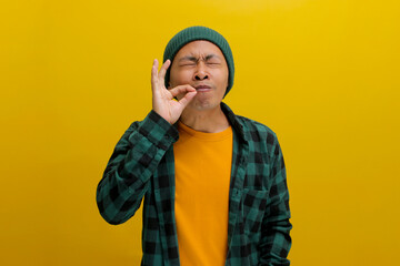Joyful young Asian man, closes his eyes and uses his fingers to make a gourmet or delicious hand gesture, expressing how enjoyable the food is while standing against a yellow background