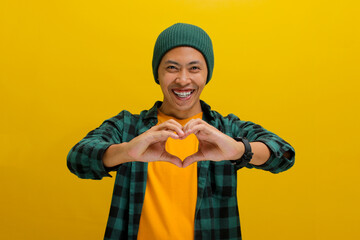 Attractive Asian man, dressed in a beanie hat and casual shirt, exudes happiness as he creates a romantic heart gesture with his hand, expressing tender feelings, standing against yellow background