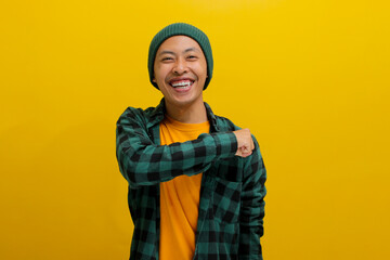 Smiling, happy young Asian man, dressed in a beanie hat and casual shirt, confidently gives a fist bump to the side while standing against a yellow background