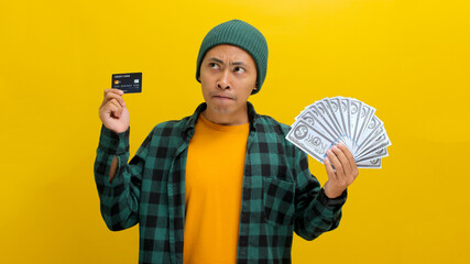 Contemplative Asian man in a beanie and casual clothes examines a credit card and cash, pondering a...