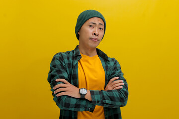 Thoughtful young Asian man, dressed in a beanie hat and casual shirt, crosses his arms, indicating uncertainty or indecision about something while standing against a yellow background