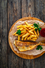 Fried sea bass served on paper with French fries and ketchup on board on wooden table 