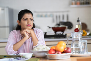 Indian woman on dining table waiting for husband or child after preparing food at home for lunch - concept of family caring, responsibility and patience