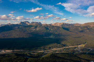 Drone view high peaks with snow under clouds sky. Forest and river below. Mountain landscape. Canada