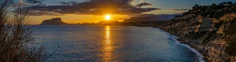 Golden sunset paints the sky as it dips below the mountainous horizon, reflecting on the...
