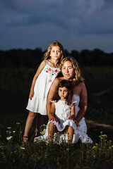 Naklejka na ściany i meble A mother and two daughters pose in white dresses at sunset. Family on vacation outside the city. Portrait of a woman with two children in nature. The concept of Mother's Day is family.