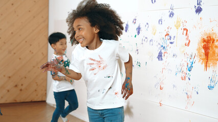 Young asian boy running to stamp colored hand print at cute african girl shirt. Student playing in...