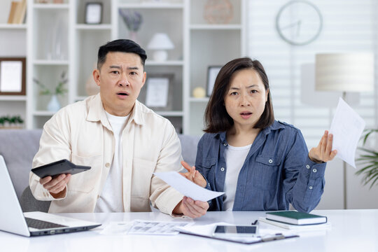 Middle-aged Asian couple managing documents and budget at home, showing confusion and concern. Laptop and paperwork spread on table.