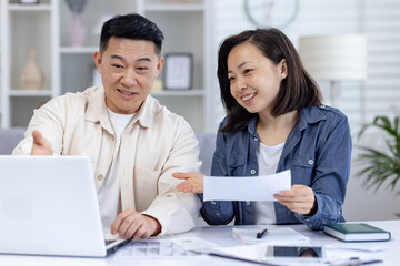 Obraz premium Asian couple engaging in household financial planning with a laptop in living room, discussing and reviewing their finances, with papers ,image captures happiness, teamwork, and digital convenience.
