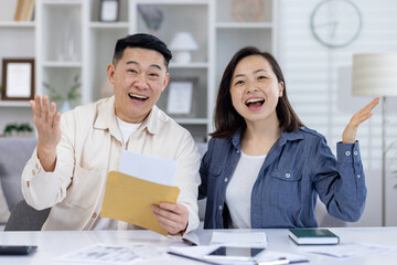 An overjoyed Asian couple in their living room, smiling and celebrating as they read some good news from a paper envelope. Their expressions reflect pure happiness and excitement.