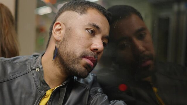 A close shot of a queer young man stressed solo traveler sitting in a moving metro train leaning on a window and looking outside. Mental health concept
