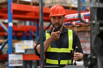 Caucasian man worker driving a forklift and using a walkie-talkie at warehouse factory container,...