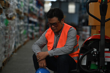 Stressed despairing mid-adult warehouse worker in a logistic business sitting at the hand truck,...