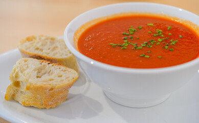A white bowl of tomato and basil soup with two slices of baguette bread on a plate.