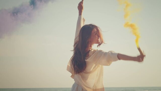 Happy asian woman with purple and yellow smoke bombs on the sea beach sunset, female activist portrait with active smoke grenades, freedom with playful dance spin slow motion