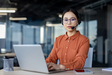 Focused young Asian woman in a modern office environment. She wears a headset and glasses, looking at her laptop screen, exhibiting professionalism and concentration.