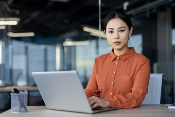 An Asian woman in a vibrant orange blouse sits seriously at her office desk, absorbed in...