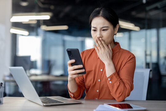 Asian woman in orange blouse looks dismayed at phone in modern office setting. Reacting to surprising bad news.