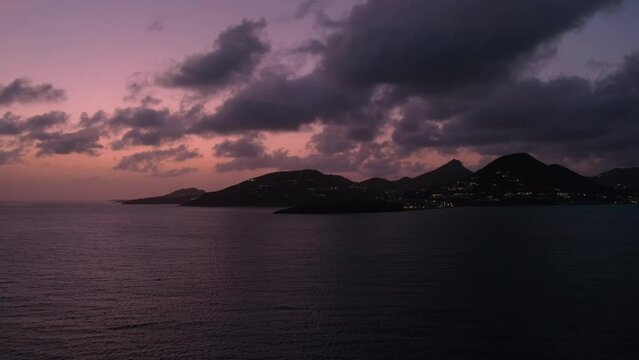 Outline of Sint Maarten in background of pink sunset sky and dusk clouds, view from cruise ship sailing in Great bay on lights of evening Philipsburg
