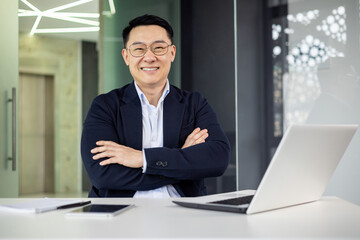 A cheerful Asian male executive stands confidently at his desk in a well-lit, contemporary office space, radiating professionalism and positivity.