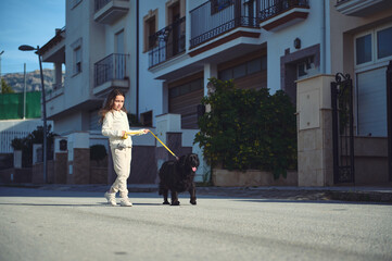 Cute child girl walking her dog on leash. Full length portrait of a happy kid in white sportswear,...