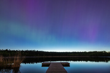 Aurora Borealis northern lights over the forest lake in Latvia on May night. Wooden pier on foreground.