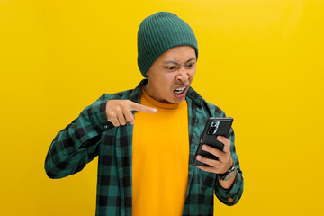 An enraged Asian man, dressed in a beanie hat and casual shirt, points at his mobile phone while visibly reacting to bad news, showing intense anger and fury, while standing against yellow background