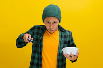 Annoyed Asian man, dressed in a beanie hat and casual shirt, switches TV channels using a remote...